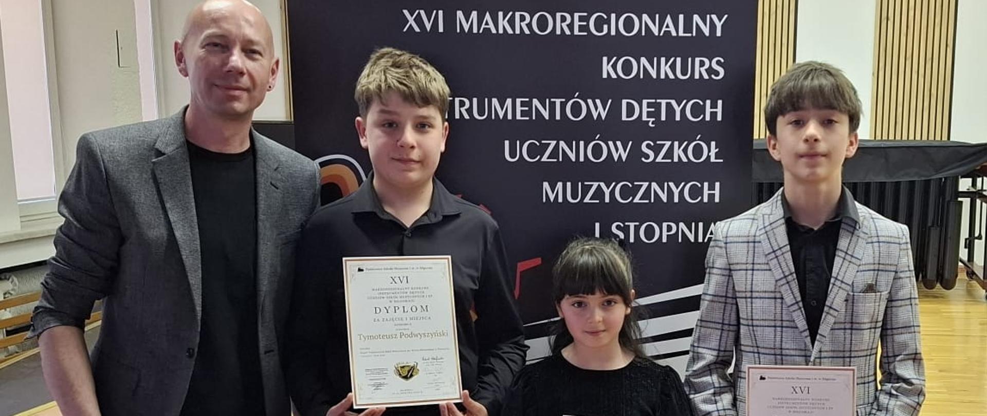 Zdjęcie pamiątkowe z XVI Makroregionalnego Konkursu Instrumentów Dętych Uczniów Szkół Muzycznych I stopnia w Biłgoraju. Na tle baneru informacyjnego Państwowej Szkoły Muzycznej I st. w Biłgoraju stoi dorosły mężczyzna oraz troje dzieci (dwóch chłopców i dziewczynka), którzy prezentują otrzymane dyplomy.
Szczegóły widoczne na zdjęciu:
Osoby: Po lewej stronie stoi mężczyzna w szarej marynarce i czarnym ubraniu. Obok niego trójka młodych wykonawców w strojach galowych, trzymających dyplomy.
Laureaci widoczni na dyplomach:
Tymoteusz Podwyszyński (dyplom za zajęcie II miejsca),
Milena Sura (dyplom za wyróżnienie),
Grzegorz Polnik (dyplom za zajęcie II miejsca).
Tło: W tle znajduje się baner konkursowy, fragment sceny oraz profesjonalne nagłośnienie. Wydarzenie odbyło się w dniach 23-24 kwietnia 2026 roku.