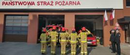 Na środku zdjęcia przed bramami garażowymi znajduje się dziesięciu strażaków jednostki ratowniczo-gaśniczej w strojach specjalnych. Po prawej stronie dwóch strażaków w strojach wyjściowych oddaje honor. 