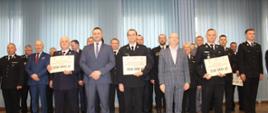Osoby w mundurach otrzymane prezentują promesy na zakup pojazdów. Strażacy OSP oraz cywile stoją w szeregu.
