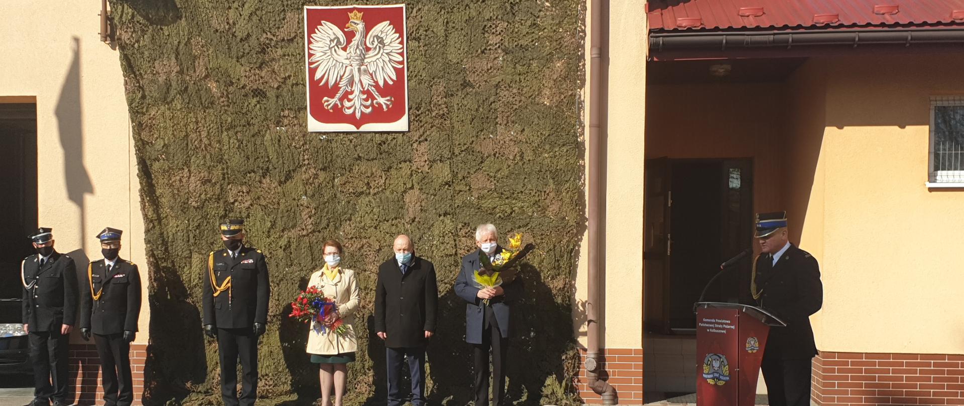 Na zdjęciu fragment budynku z zawieszoną maskownicą oraz wiszącym na niej godłem. Na tle strażacy w umundurowaniu galowym oraz osoby cywilne. Jeden strażak przemawia przy mównicy. 