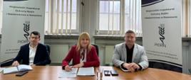 Trzy osoby siedzą przy stole konferencyjnym w sali biurowej. Na środku kobieta podpisuje dokumenty, po jej bokach siedzą dwaj mężczyźni. W tle okna z pionowymi żaluzjami oraz roll-upy Wojewódzkiego Inspektoratu Ochrony Roślin i Nasiennictwa w Rzeszowie.