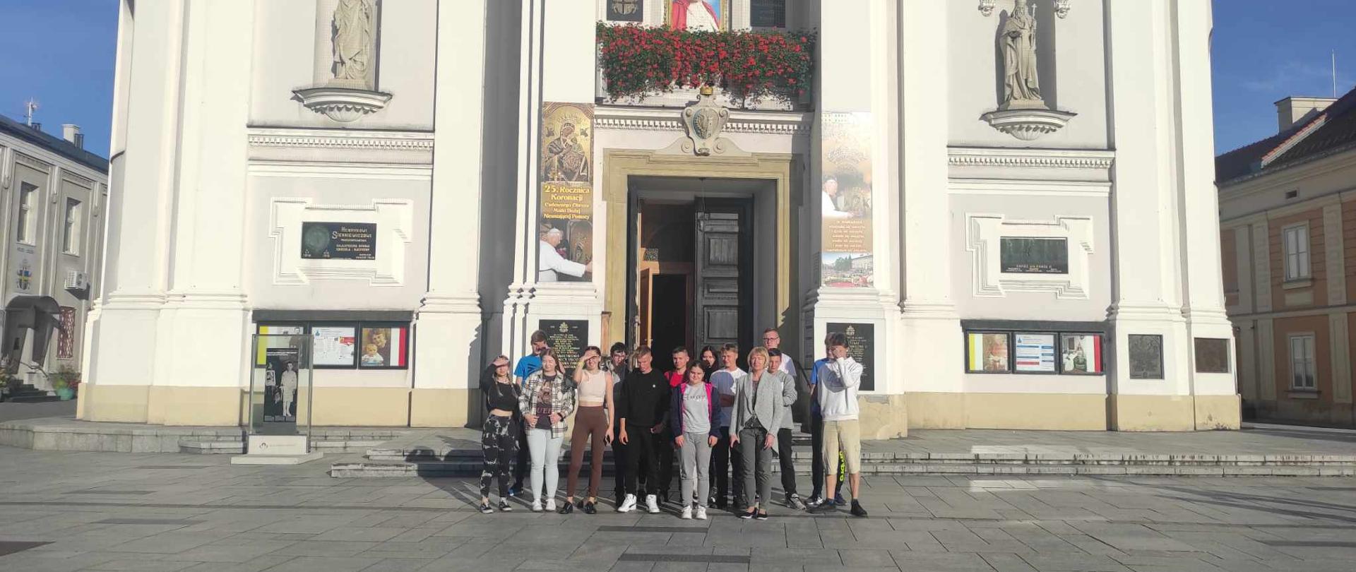 Grupa osób stoi przed wejściem do białego kościoła o klasycystycznej fasadzie, udekorowanego wizerunkami i flagami papieskimi.