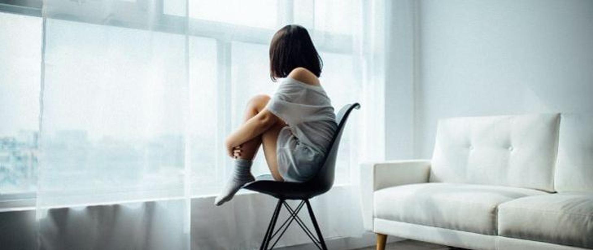 Samotna dziewczynka z podkulonymi nogami siędząca na krześle patrząca w okno 