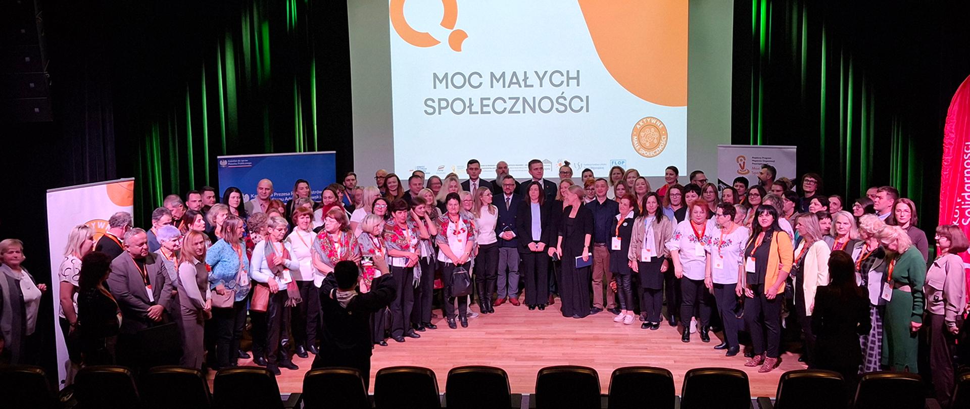 Szeroki kadr, zdjęcie grupowe. Uczestnicy konferencji pozują do wspólnego zdjęcia, stojąc razem na scenie. Za nimi widoczny ekran, na którym wyświetlona jest grafika z tytułem: Moc Małych Społeczności. Za uczestnikami, po obu stronach ekranu widoczne są także fragmenty banerów promocyjnych.