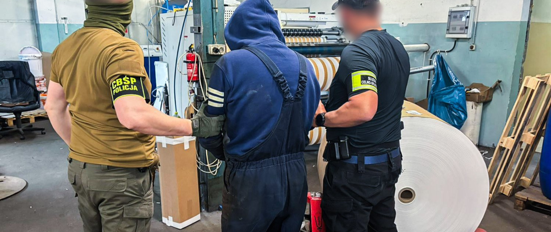 Dwóch funkcjonariuszy w oznaczeniach CBŚP i Służby Celno-Skarbowej trzyma osobę w roboczym stroju przy maszynie z dużą rolą materiału w hali przemysłowej.