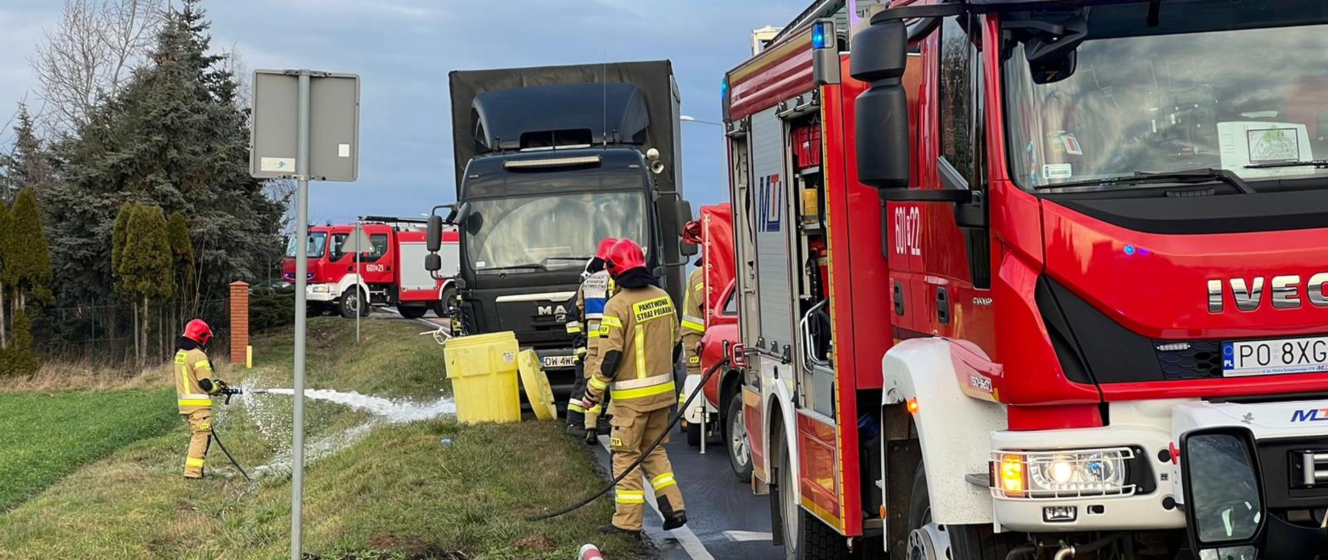 Na zdjęciu czerwone samochody strażackie oraz samochód ciężarowy koloru czarnego biorący udział w zdarzeniu, strażak podający prąd piany pod uszkodzony samochód ciężarowy.