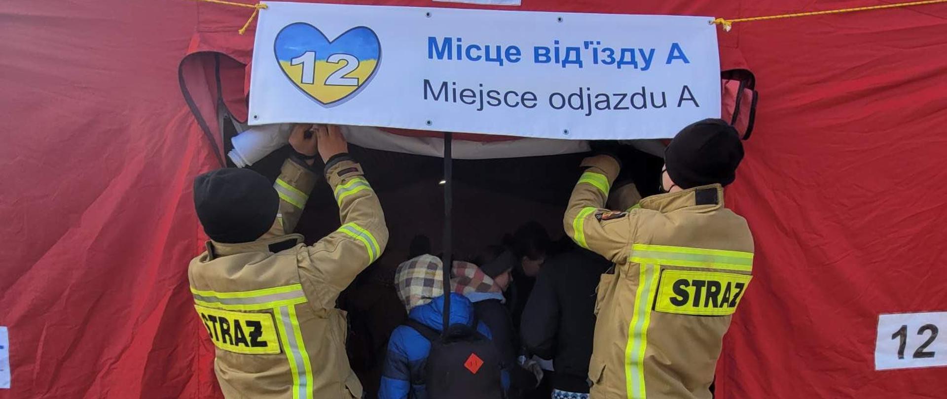 Na zdjęciu widać dwóch strażaków PSP w ubraniu specjalnym przypinających baner na namiocie z oznaczeniem miejsca odjazdu dla uchodźców. W namiocie widać dzieci. 
