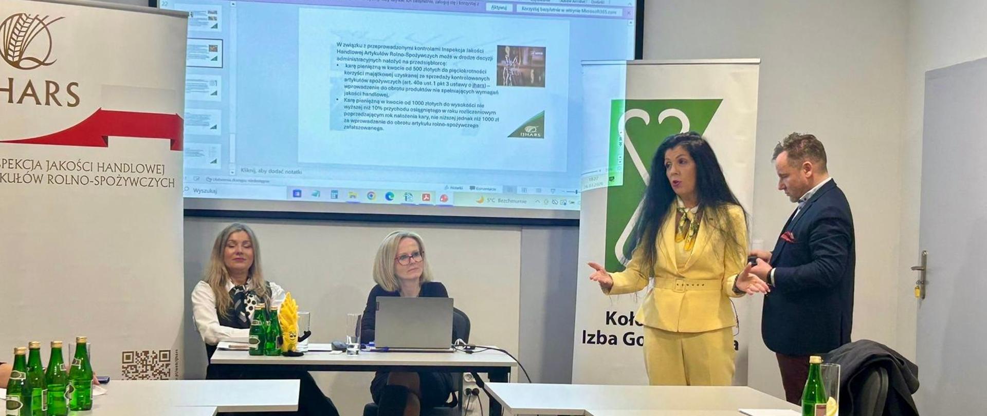 Inspektorki siedzą przy biurku i prezentują materiał, natomiast przy tablicy stoi Anna Kusy-Łowicka, która omawia zagadnienia – w tle widoczna jest wyświetlana prezentacja.