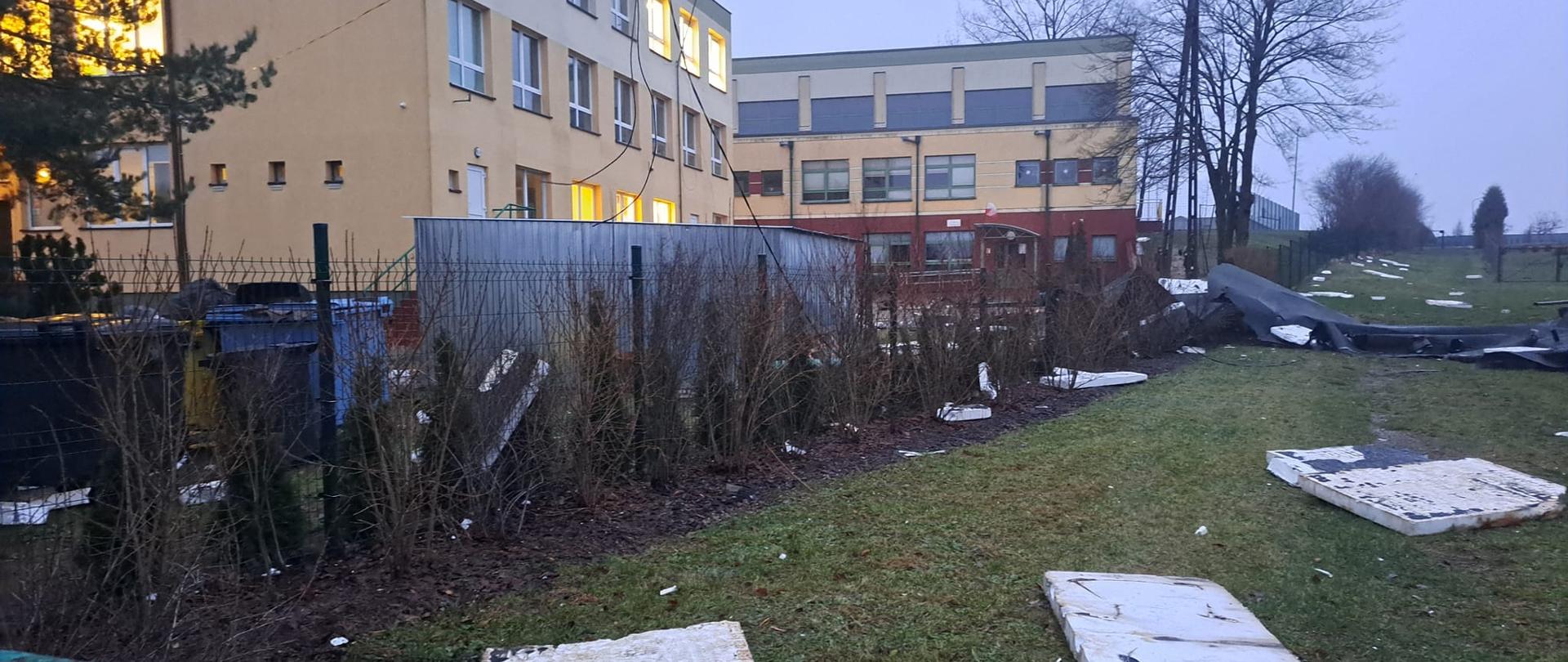 Porozrzucany styropian leży na trawie niedaleko znajduje się ogrodzenie garaż oraz budynek szkoły.