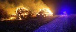 Zdjęcie przedstawia palące się baloty siana. Pora nocna. Na zdjęciu widać również samochód strażacki. 