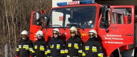 Widok z przodu. Pięciu strażaków OSP Skawinki stoi w ubraniach specjalnych przed samochodem pożarniczym przed garażem OSP. 
