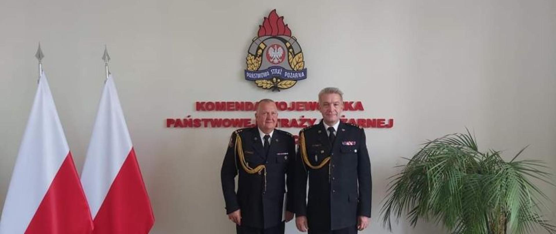 W centralnym punkcie zdjęcia stoi dwóch strażaków. Zastępca Komendanta Wojewódzkiego oraz przejmujący obowiązki Komendanta Powiatowego Zastępca KP PSP Białogard. Po lewej stronie zdjęcia flagi Polski. W tle logo PSP a po prawej zielony kwiat