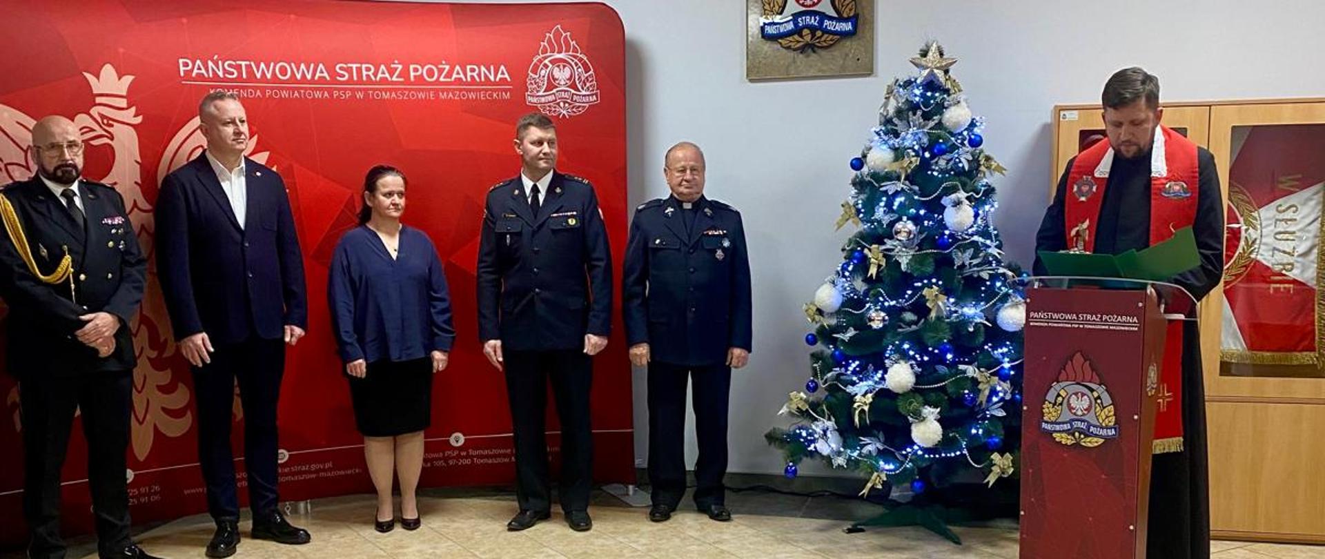 Zaproszeni goście stoją obok siebie w tle czerwony baner PSP oraz choinka świąteczna. 
