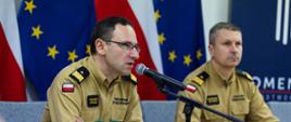 Strażak PSP w żółtym mundurze służbowym mówi do mikrofonu podczas konferencji prasowej; obok niego siedzi inny funkcjonariusz w podobnym umundurowaniu. W tle widoczne są flagi Polski i Unii Europejskiej.