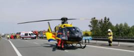 Zdjęcie przedstawia helikopter lotniczego pogotowia ratunkowego, który stoi na pasie autostrady A2. Z tylu widać samochody strażackie, karetkę pogotowia ratunkowego oraz strażaków. 