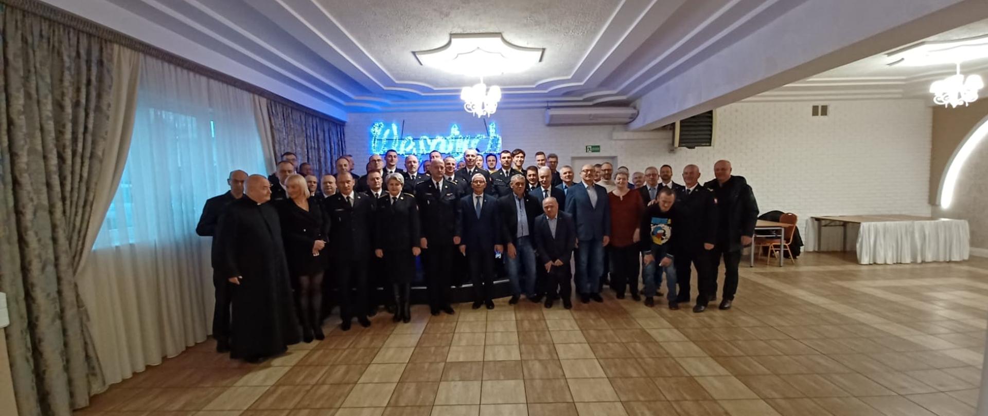 Wszyscy uczestnicy spotkania stoją w zwartej grupie i pozują do zdjęcia. W tle podświetlany napis "Wesołych świąt"