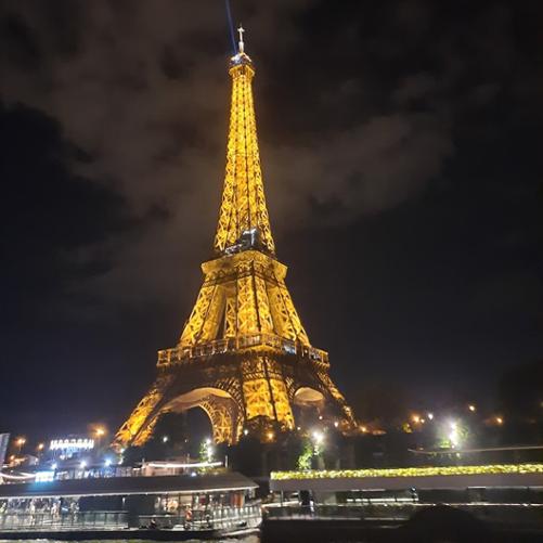 Zdjęcie wieży Eiffla nocą. Przed wieżą z lewej i prawej strony na rzece 2 statki wycieczkowe. W tle za wieżą widoczne chmury.