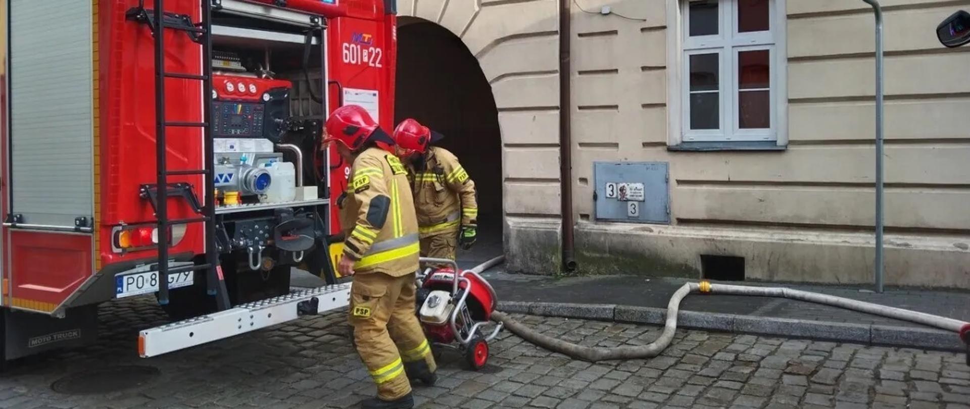 Na zdjęciu samochód ratowniczo - gaśniczy przy budynku z rozwiniętą linią wężową za pojazdem stoją ratownicy ubrani w ubrania specjalne wkładają wentylator na pojazd 