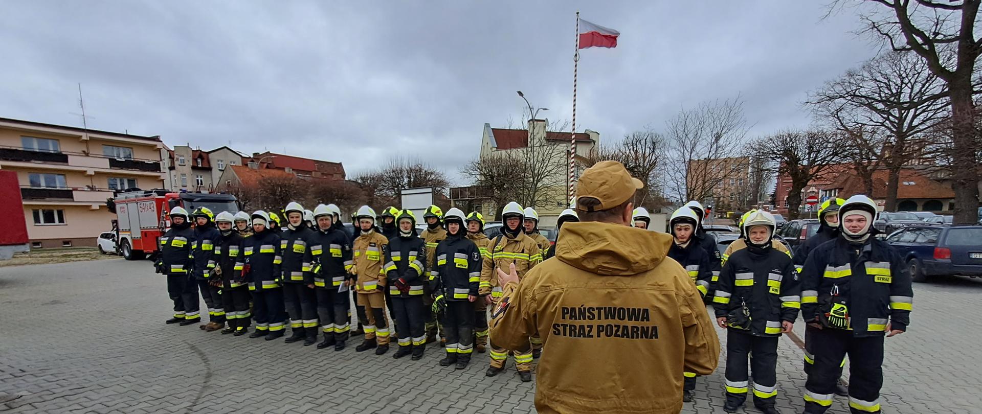 Plac Komedy PSP w Tczewie. Na pierwszym planie stoi instruktor tyłem do zdjęcia, na plecach napis PAŃSTWOWA STRAŻ POŻARNA. Mówi do stojących przed nimi kandydatów na strażaków ratowników ustawionych w dwuszeregu. W tle samochód pożarniczy i maszt z powiewającą flagą Polski.