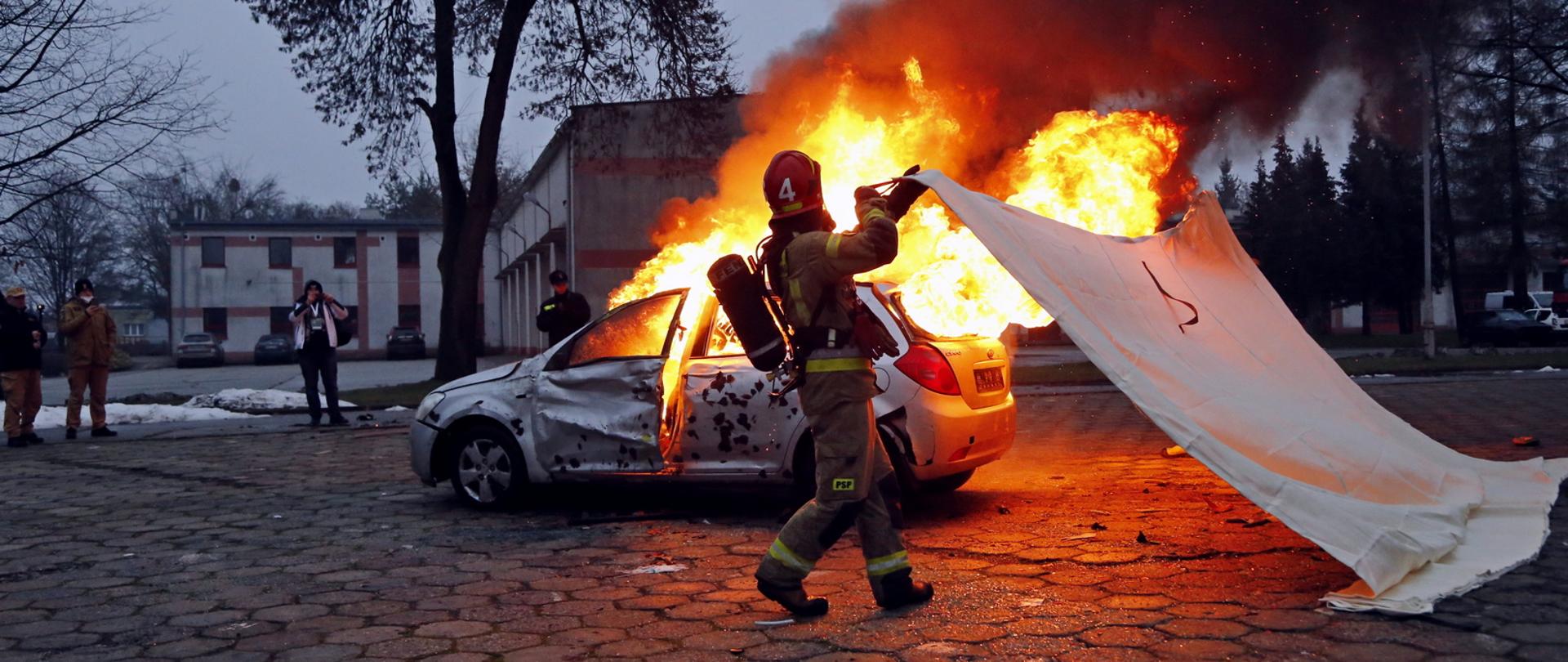 Poligon CS PSP, widoczny płonący wrak samochodu osobowego do którego zbliża się dwóch strażaków trzymając rozciągniętą płachtę
