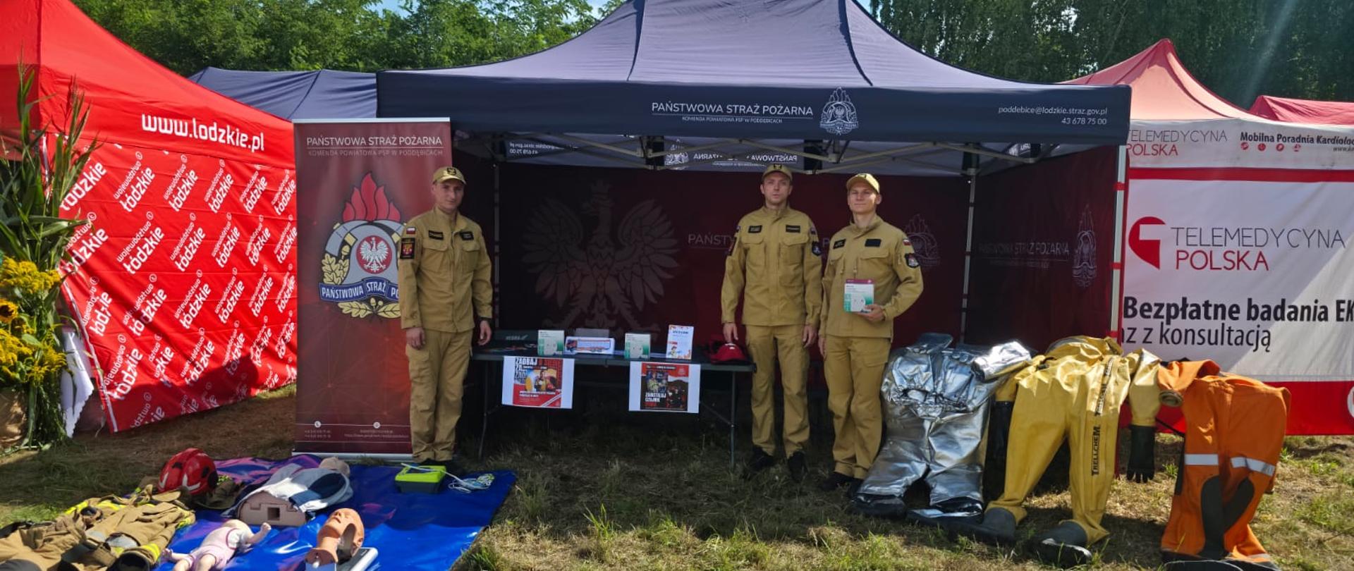 trzej strażacy stoją przed namiotem z napisem państwowa straż pożarna jeden z nich trzyma czujkę dymu . obok na stoliku leża różne stroje których używają strażacy podczas działań ratowniczych
