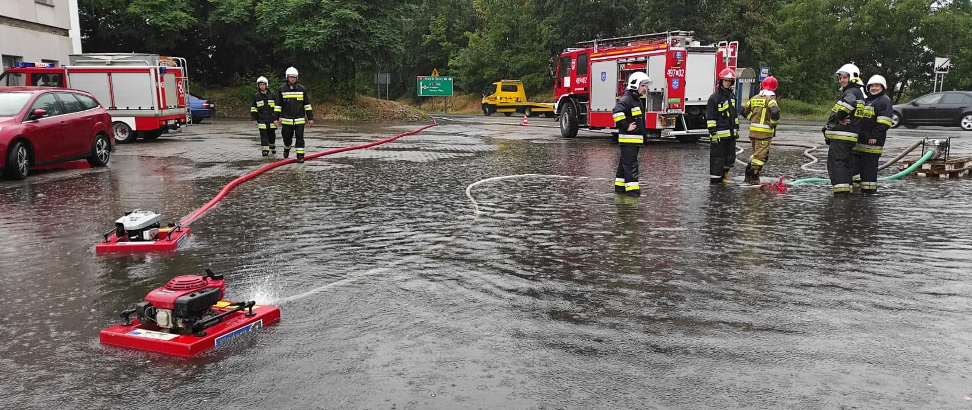 Strazacy obsługujący pompę pływającą. Strażak w umundurowaniu bojowym. Zalany plac stacji paliw. W tle budynek i pojazd pożarniczy.