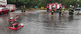 Strażacy obsługujący pompy pływającą. Strażacy w umundurowaniu bojowym. Zalany plac stacji paliw. W tle budynek i pojazd pożarniczy.