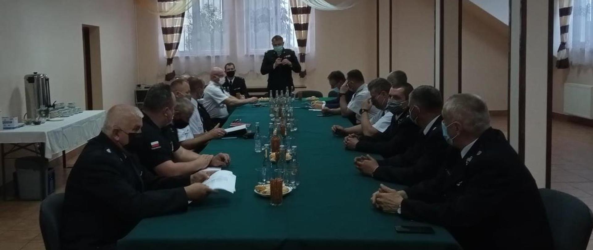 
W pomieszczeniu przy stole siedzą strażacy. Na końcu stołu stoi strażak w mundurze generała, który trzyma w ręku mikrofon i przemawia. 