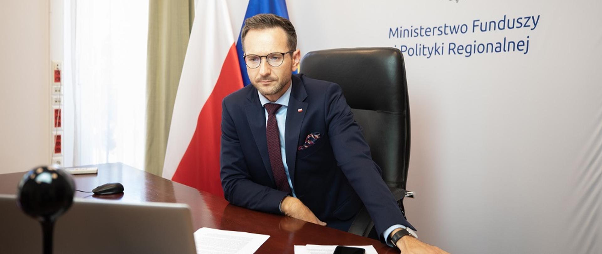 Waldemar Buda siedzi za biurkiem, przed nim laptop, za nim fagi Polski i UE oraz tło z logo MFiPR.