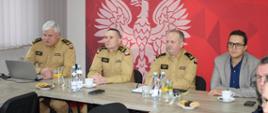 Na pierwszym planie przy podłuznym stole siedzi 4 panów (od lewej): w żółtym ubraniu służbowym Zastępca Komendanta Powiatowego PSP st. bryg. Paweł Borek, w żółtym ubraniu służbowym Świętokrzyski Komendant Wojewódzki PSP st. bryg. Marcin Charuba, w żółtym ubraniu służbowym Komendant Powiatowy PSP mł. bryg. Sławomir Zięba, na końcu siedzi osoba w okularach, szarej marynarce i granatowej koszuli Starosta Starachowicki Piotr Babicki. Za plecami mają czerwony baner z białym godłem Polski.