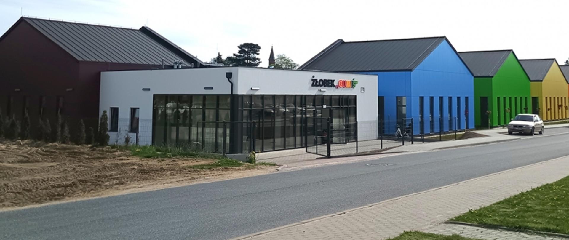 Budynek, w którym znajduje się żłobek. Ma on prosta bryłę i przeszkolone ściany. Za nim usytuowane są 3 budynki ze spadzistym dachem. Każdy z nich jest w innym kolorze.