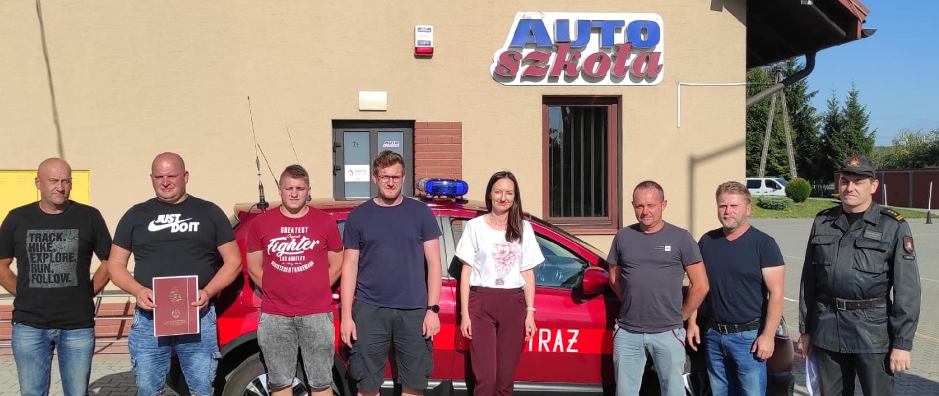 Zdjęcie przedstawia Komendanta Powiatowego PSP w Brzozowie, kierownika ośrodka szkolenia kierowców wraz z kursantami, którzy rozpoczynają szkolenie. Osoby te znajdują się przed budynkiem Auto-Szkoły w Brzozowie, w tle samochód Państwowej Straży Pożarnej
