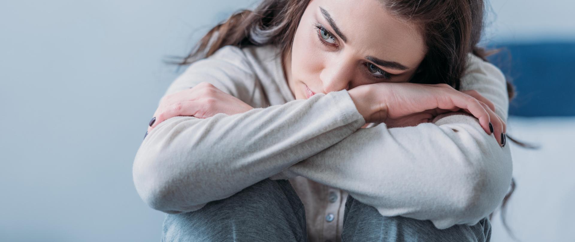 Na zdjęciu jest kobieta, która ma ręce oparte o podkulone nogi. Głowę opiera o ręce. Na jej twarzy widoczny jest smutek. Tło jest w odcieniach siwego oraz niebieskiego.