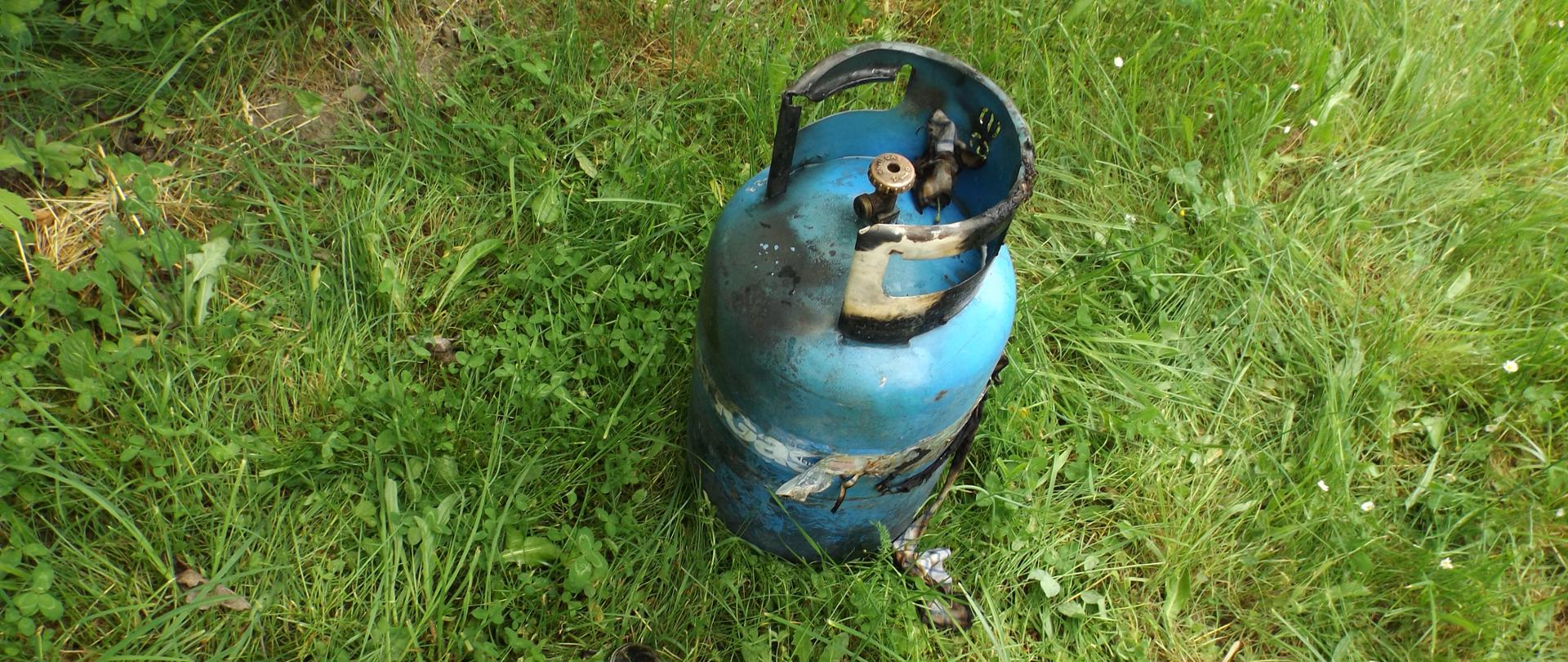 Zdjęcie przedstawia butlę 11kg stojącą na trawie. Widać ślady okopcenia świadczące o pożarze butli.