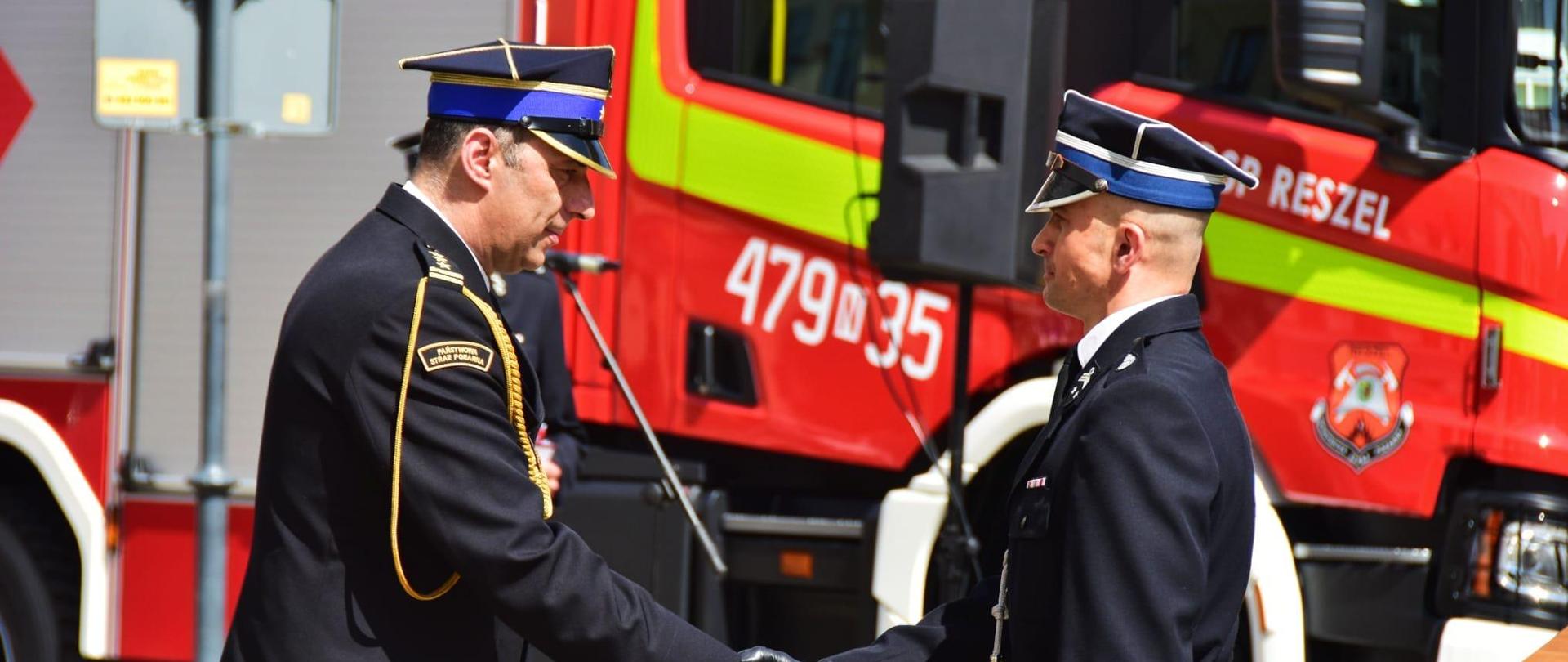 Na zdjęciu widać dwóch strażaków w uścisku dłoni. Funkcjonariusze ubrani w mundury wyjściowe. Za nimi można zauważyć pojazd strażacki.