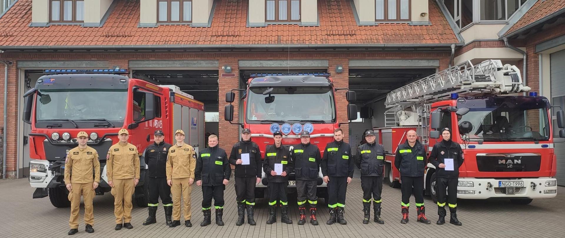 Zdjęcie przedstawia uczestników szkolenia kierującego działaniem ratowniczym dla strażaka ratownika Ochotniczych Straży Pożarnych (dowódcy OSP), realizowanego w Komedzie Powiatowej Państwowej Straży Pożarnej w Gołdapi, którzy wspólnie z komisją egzaminacyjną, stoją na tle pojazdów pożarniczych zlokalizowanych w garażach strażnicy