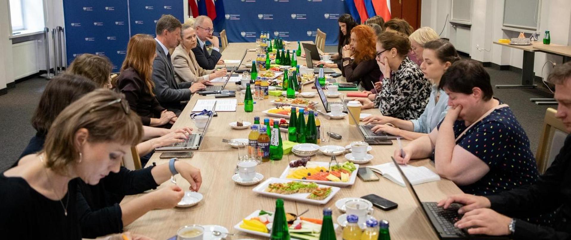 Spotkanie przy stole konferencyjnym; uczestnicy pracują na laptopach, na stole woda, kawa i przekąski, w tle ścianka z logo i flagi.