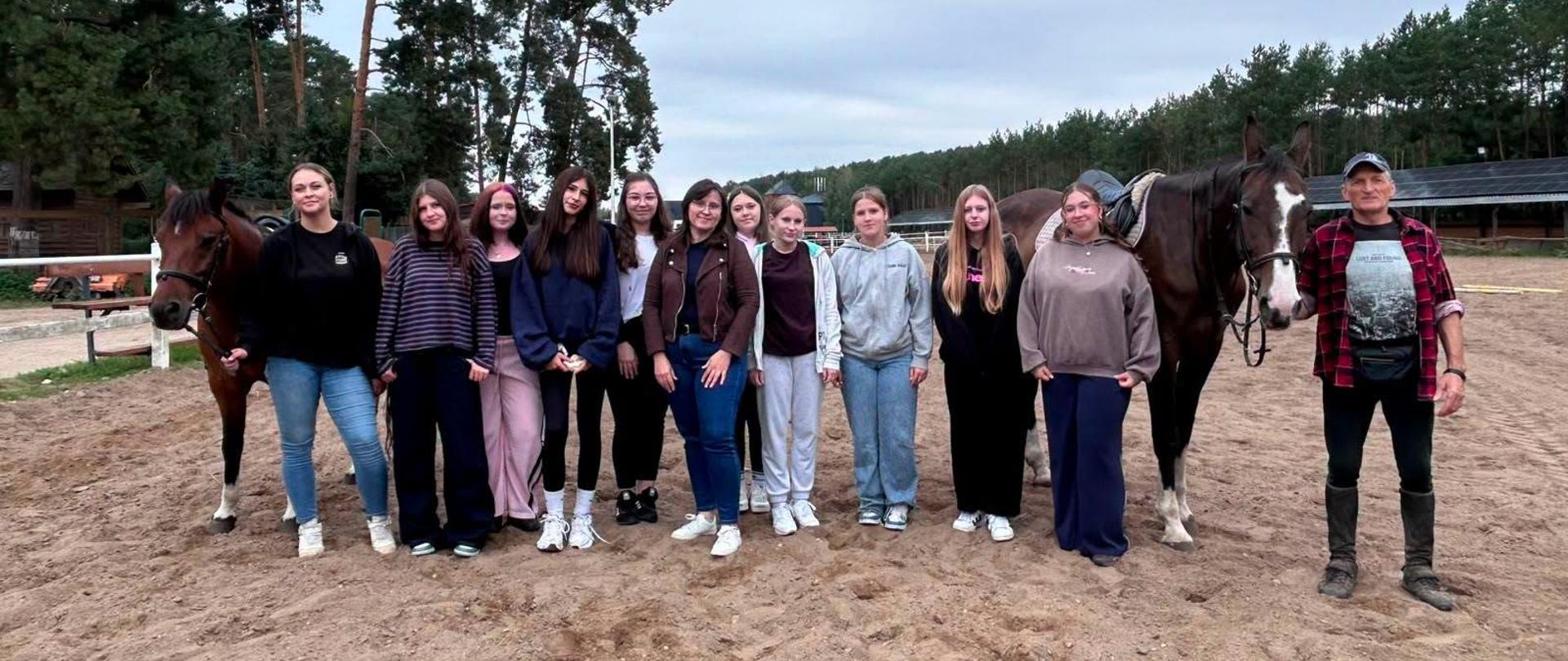 Grupa uczennic oraz opiekun pozują do pamiątkowego zdjęcia na piaszczystym wybiegu w otoczeniu koni.