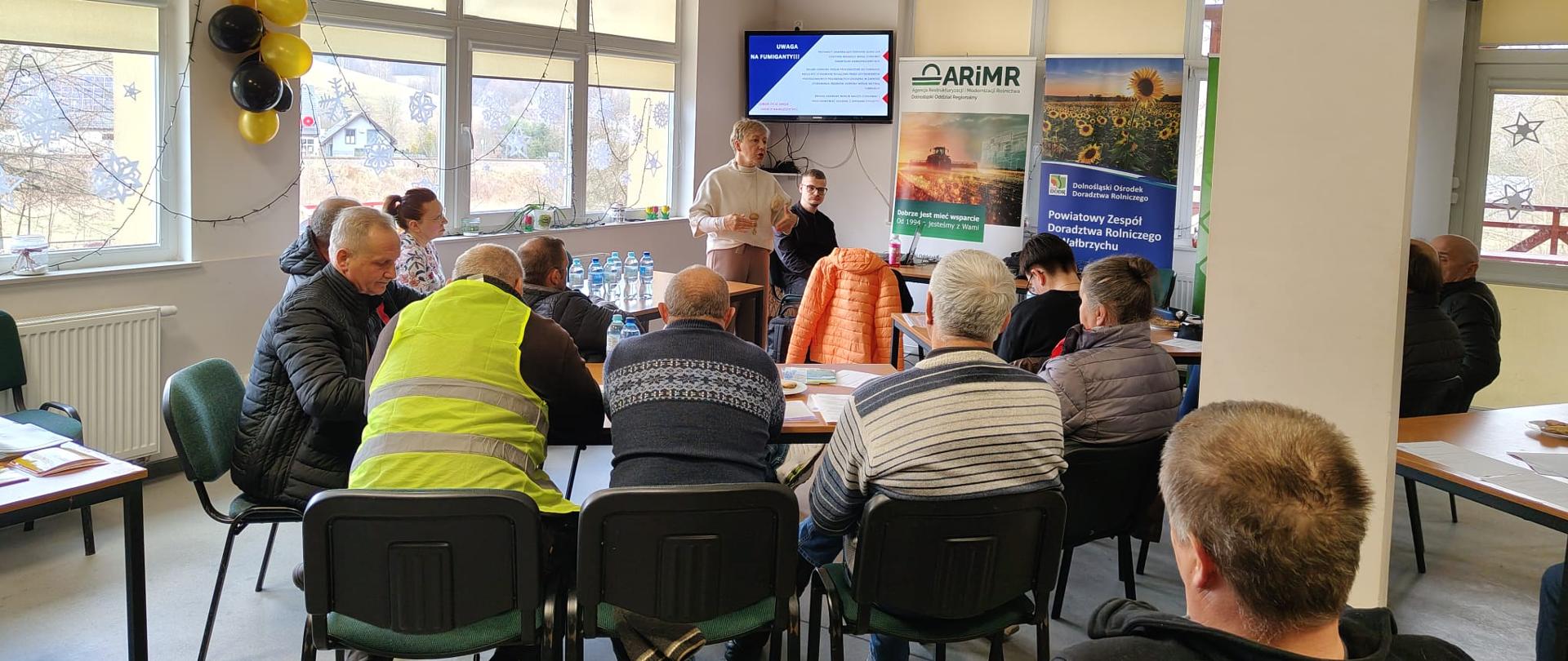 Rolnicy siedzą przy stole w sali konferencyjnej, w tle widoczny wykładowca 