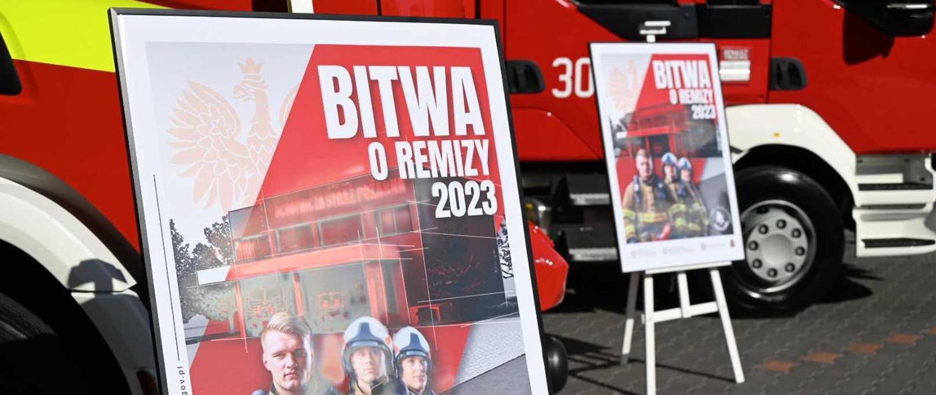 Dwa samochody ciężarowe straży pożarnej barwy czerwonej na których tle stoją na stojaku dwa banery reklamowe z napisem bitwa o remizy 2023
