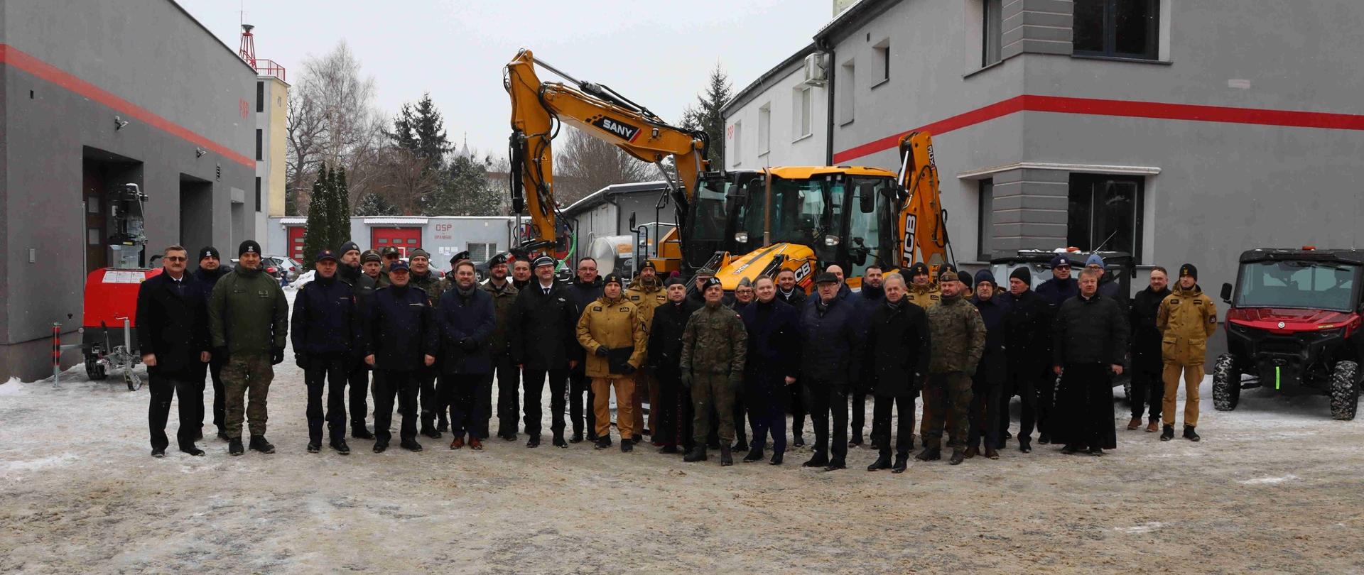 Na zdjęciu duża grupa uczestników spotkania. Pozują do zdjęcia na tle maszyn budowlanych i strażackich. Warunki pogodowe zimowe.
