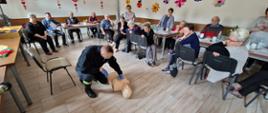Zdjęcie przedstawiające seniorów przyglądających się pokazowi. Seniorzy siedzą przy stołach a strażacy na środku sali przeprowadzają pokaz.