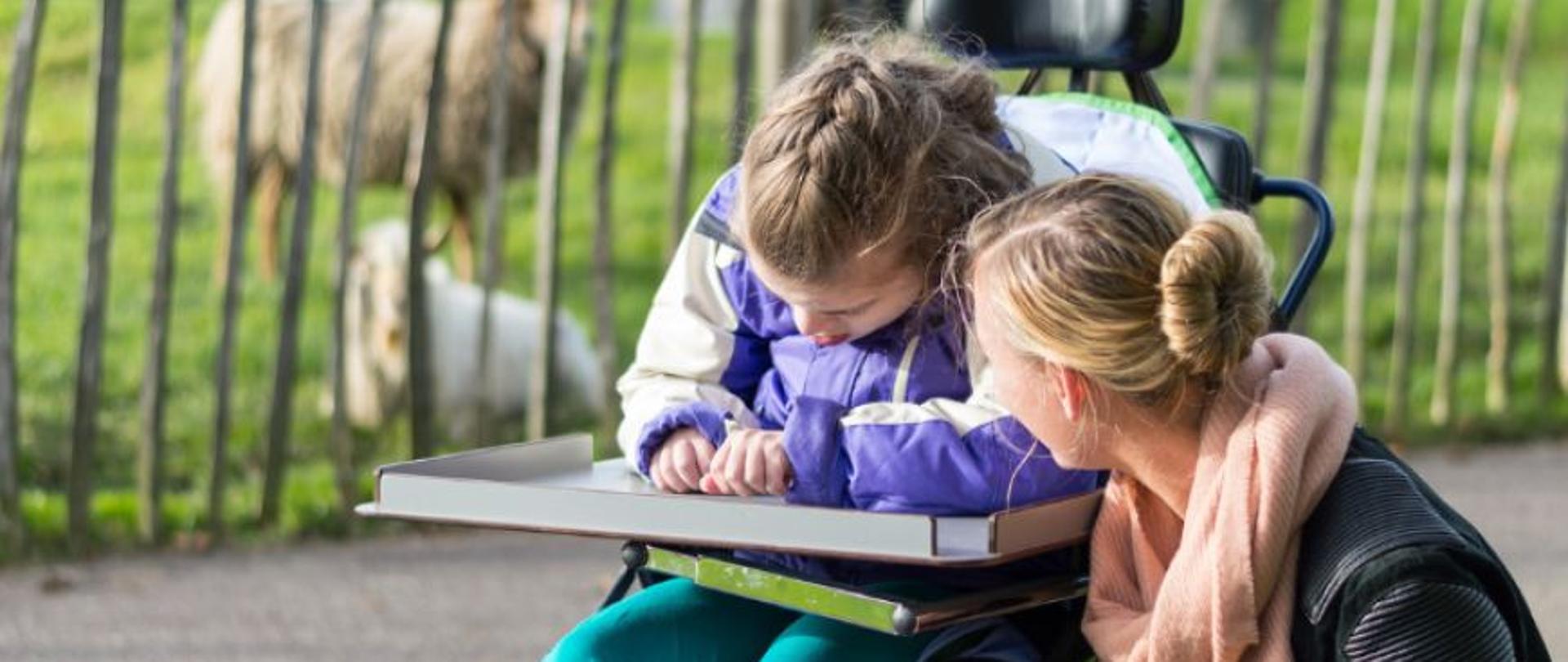 Dziewczynka na wózku inwalidzkim, obok kuca kobieta , w tle płot a za nim owieczki