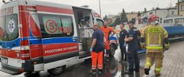 Na zdjęciu widać uszkodzony pojazd ciężarowy. Po lewej pojazd Państwowego Ratownictwa Medycznego, który posiada sygnalizacją ostrzegawczą na dachu. W pobliżu funkcjonariusz Policji, PSP oraz członkowie Zespołu Ratownictwa Medycznego. Asfalt mokry od opadów deszczu. Niebo zachmurzone. W tle domy jednorodzinne.