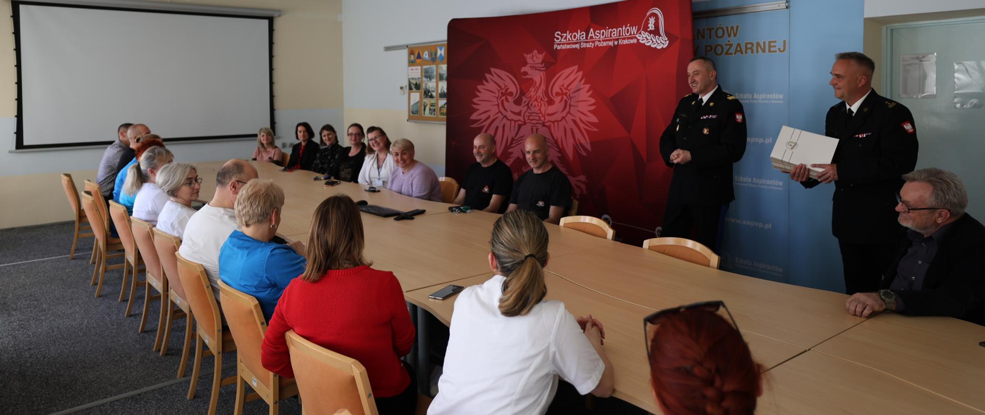 Zdjęcie wykonane w długiej jasno oświetlonej sali. Na jej środku widoczny długi stół w kolorze drewna. Na ścianie naprzeciwko fotografa oficialne tablo Państwowej Straży Pożarnej w kolorze czerwonym. Na nim białe godło polski z napisem Szkoła Aspirantów PSP w Krakowie. Wokół stołu siedzą osoby w ubraniach cywilnych , którzy byli uczestnikami szkolenia. Po prawej stołu naprzeciwko fotografa stoją dwaj funkcjonariusze w galowych mundurach Państwowej Straży Pożarnej jeden z nich trzyma w rękach białą teczkę z dyplomem. Atmosfera jest swobodna osoby mają uśmiechy na twarzach. 