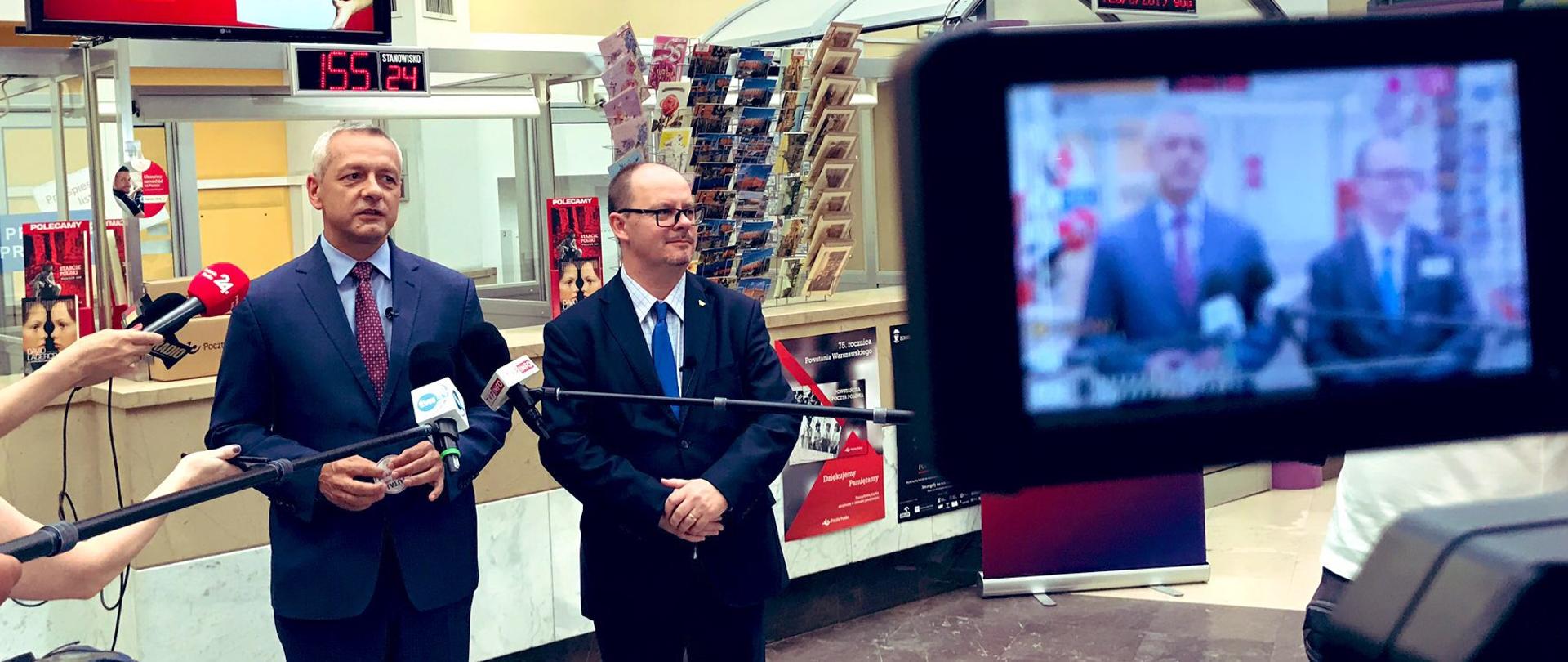 Minister cyfryzacji Marek Zagórski i prezes Poczty Polskiej Przemysław Sypniewski stoją przed okienkami pocztowymi