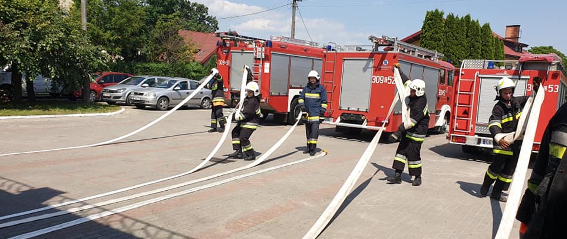 Strażacy w granatowych ubraniach specjalnych ćwiczący rozwijanie węży na placu z kostki, w tle 3 wozy strażackie