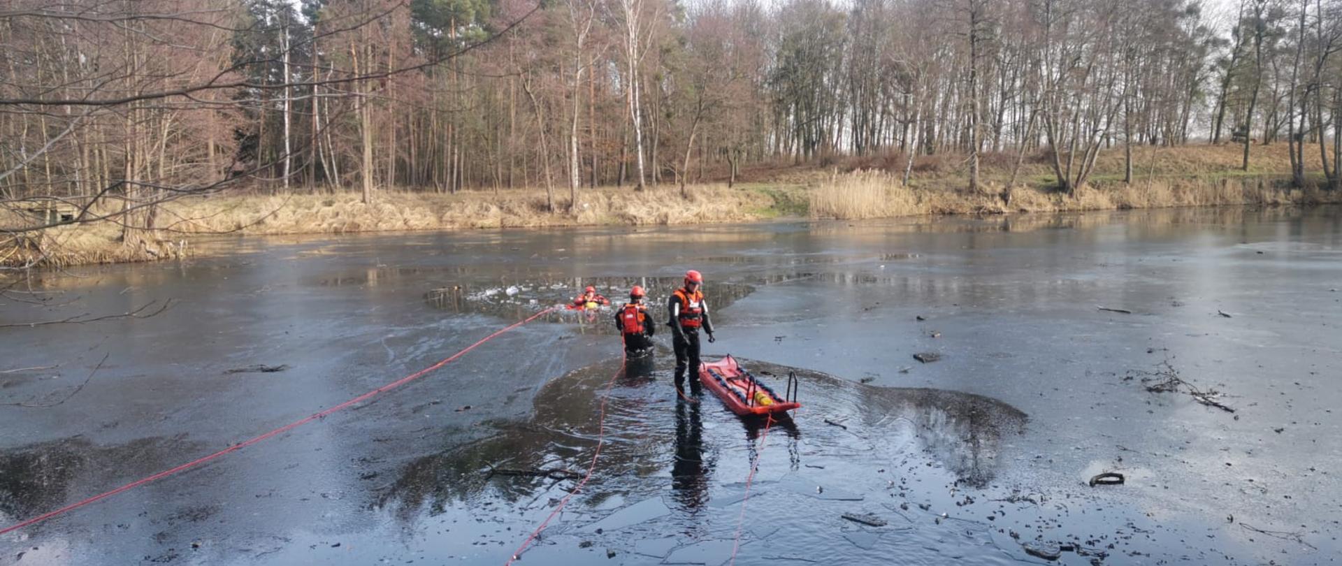 Ratownicy w ubraniach wypornościowych przeprowadzają podejście z deską lodową do osoby, pod którą załamał się lód.
