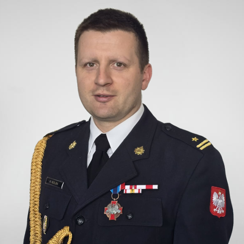 Komendant PSP na zdjęciu w mundurze wyjściowym. Zdjęcie obejmuje popiersie Komendanta. Na mundurze widoczne odznaczenia pożarnicze oraz imiennik