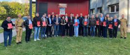 Grupa kilkunastu osób pozuje do pamiątkowego zdjęcia na tle czerwonego budynku z napisem „Państwowa Straż Pożarna”. Uczestnicy spotkania stoją na trawniku przed budynkiem, niektórzy z nich trzymają w dłoniach czerwone teczki z godłem Polski. Wśród zgromadzonych widać osoby w strojach cywilnych oraz w mundurach strażackich.